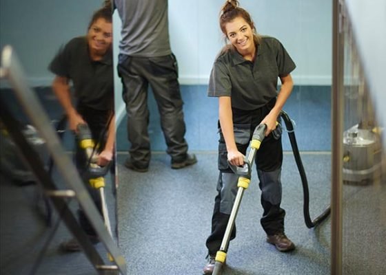 a female cleaning contractor steam cleans an office carpet in a empty office in between tenants. In the background a male colleague is polishing the glass partition offices .